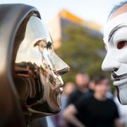 En man med Guy Fawkes-mask kollar på statyn av bitcoins grundare Satoshi Nakamoto 