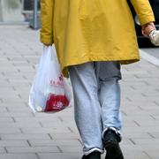 En vuxen person med gul jacka och en plastpåse i ena handen håller ett barn i andra handen när de promenerar iväg hand i hand på trottoaren. 