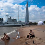 Två män som badar framför ett militärfartyg och statliga oljejätten Gazproms huvudkvarter i Sankt Petersburg. 