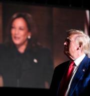 Donald Trump talar framför en skärm som visar Kamala Harris under ett kampanjmöte. 