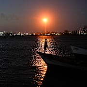 En pojke står på en fiskebåt med Cardón-raffinaderiet i bakgrunden i Punta Cardón i Venezuela.