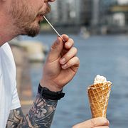 En man med tatuerad arm sitter på kajen och äter smältande glass i strut. Illustrationsbild.