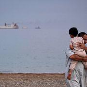 En man bär ett barn på en strand medan oljetankers och lastfartyg köar i Hormuzsundet utanför Förenade arabemiraten.